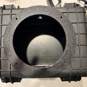 May include: A black rectangular hard-shell storage case featuring a grid-patterned exterior and a large circular cutout in the center. The circular opening is framed by a textured, dark gray mounting ring with four small screw holes for installation.