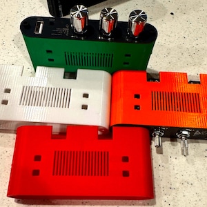 May include: Four rectangular electronic amplifier enclosures in green, white, orange, and red. Each unit features a black top panel with three metallic control knobs, a USB port, and an auxiliary input. The front faces have a central slotted vent.