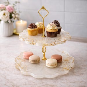 Könnte beinhalten: Ein zweistufiges Serviertablett mit goldenem Griff, das Cupcakes und Macarons enthält. Das Tablett hat ein weißes und goldenes Design. Die Cupcakes haben Schokoladen- und Vanille-Frosting. Die Macarons sind rosa, beige und braun.
