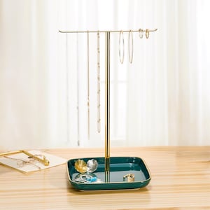 May include: A gold-colored jewelry stand with a square, teal-colored base and a vertical post. The stand holds necklaces, earrings, and rings. The base has a gold trim. The jewelry stand is on a wooden surface.