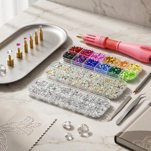 Könnte beinhalten: Bastelbedarf zum Verzieren. Enthält einen pinkfarbenen Heißklebeapplikator, Schalen mit bunten Strasssteinen, eine silberne Schale mit goldfarbenen Werkzeugen und Edelsteinen sowie ein Notizbuch.