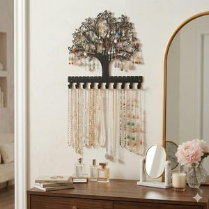 Könnte beinhalten: Schwarzer Schmuck-Organizer in Baumform aus Metall mit hängenden Halsketten und Ohrringen. Das Baumdesign hat Blätter und Äste zur Präsentation von Schmuck. Der Organizer ist an einer weißen Wand über einer Holzkommode montiert.