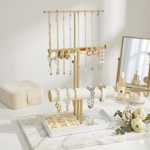 May include: A gold-toned jewelry organizer with multiple levels for necklaces, bracelets, rings, and watches. The organizer is filled with various necklaces, bracelets, and rings. A small, beige jewelry box and a mirror are also visible.