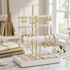 Op de afbeelding: Een goudkleurige sieradenorganizer met meerdere niveaus voor kettingen, oorbellen, armbanden en ringen. De organizer heeft een witte basis en een goudkleurig metalen frame. Diverse goudkleurige sieraden worden getoond.