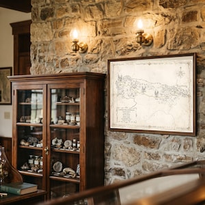 May include: A framed, rectangular, off-white map with black ink illustrations and text titled Camino de Santiago, featuring a compass rose in the top right corner, displayed on a textured stone wall above a wooden cabinet with glass doors.