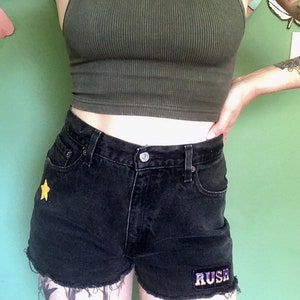 May include: Black denim shorts with a yellow star and a "RUSH" patch on the front. The shorts have a frayed hem.
