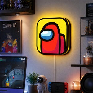 Pode incluir: Uma luz LED de parede com o tema Among Us. A luz apresenta o personagem vermelho do jogo em um fundo amarelo. A luz tem uma borda preta e está conectada a uma tomada.