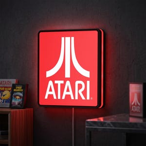 May include: A square, red Atari light-up sign with the Atari logo in white. The sign is mounted on a dark gray wall. The sign is lit with a red glow. A small Atari box is on a nearby table.
