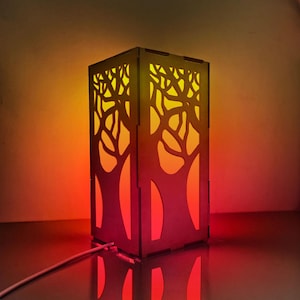 Puede incluir: Lámpara rectangular de madera con un diseño de ramas de árbol cortado en los lados. La lámpara emite un brillo cálido y colorido, que va del rojo en la base al amarillo en la parte superior. Un cable blanco sale de la base.