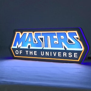 Może przedstawiać: Prostokątny podświetlany znak ścienny LED z napisem MASTERS OF THE UNIVERSE pogrubioną niebieską czcionką z białym obrysem. Znak jest otoczony świecącą pomarańczową ramką i emituje niebieskie światło, z czarnym kablem zasilającym z boku.