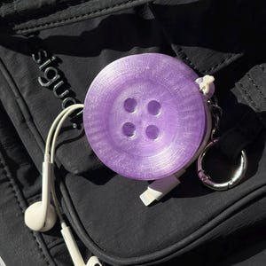 Puede incluir: Un estuche redondo de plástico morado translúcido con forma de botón de cuatro agujeros. El estuche cuenta con un llavero de metal plateado y un cable de carga blanco con conector lightning. Se incluyen auriculares con cable de color blanco.