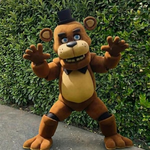 May include: A brown bear costume with a black top hat and bow tie. The bear has a tan belly and blue eyes. The costume is standing in front of a green hedge.