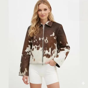 May include: A brown and white cowhide jacket with a zipper closure. The jacket has a collared neckline and long sleeves. The model is wearing white shorts.