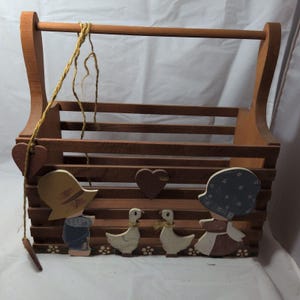 May include: A brown wooden slatted basket with a top handle, featuring painted cutouts of two children and two geese. A small wooden heart is attached to the side with a yellow string, and a larger heart is centered on the front slats.