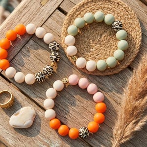 May include: Three beaded bracelets featuring round silicone beads in orange, white, sage green, and light pink. Each bracelet includes patterned beads with a black and white leopard print and a small gold-toned smiley face charm accent.