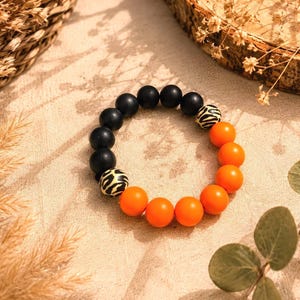 May include: A stretch bracelet featuring a series of round beads in matte black and vibrant orange. The design includes two accent beads with a black and beige animal print pattern, arranged in a circular formation on a neutral fabric surface.