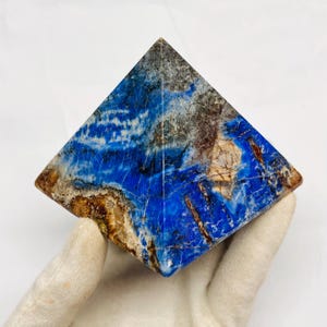 Può includere: Una piramide in pietra lucida caratterizzata da un colore di base blu vibrante con venature naturali complesse nelle tonalità del bianco, grigio e marrone. La superficie presenta una finitura liscia e lucida che esalta i motivi minerali.