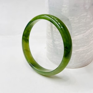 May include: A smooth, polished bangle bracelet crafted from translucent green stone. The circular accessory features natural variations in color, ranging from light lime to deep forest green, with a glossy finish that reflects light across its surface.