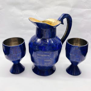 Può includere: Un set composto da una brocca e due calici abbinati, tutti caratterizzati da un esterno a mosaico blu intenso con segmenti di pietra visibili. La brocca presenta un manico curvo e un interno dorato, mentre i calici hanno interni metallici.