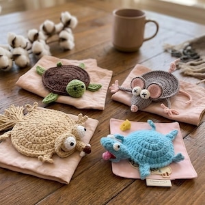 May include: Four handmade crochet animal coasters: a brown turtle, a gray mouse, a tan creature, and a blue creature. Each coaster is placed on a pink square cloth. A brown mug and cotton sprigs are in the background.