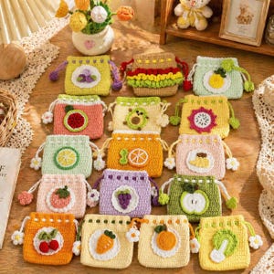 Peut inclure: Une collection de pochettes à cordon colorées, crochetées, chacune ornée d'un motif de fruit ou d'aliment différent. Les pochettes sont de différentes couleurs, dont le jaune, le rose, le vert et l'orange, et sont disposées sur une surface en bois.