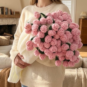 Puede incluir: Un gran ramo de flores rosas tejidas a ganchillo con tallos y hojas verdes. Las flores están muy juntas y envueltas en una tela blanca tejida a ganchillo, creando una apariencia suave y texturizada. El ramo lo sostiene una persona que lleva un suéter color crema.
