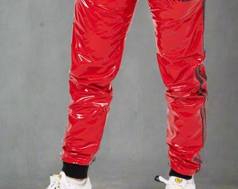 May include: Bright red glossy PVC track pants featuring a black vertical stripe down each leg, black ribbed ankle cuffs, and the text PVCUP printed in black on the upper left thigh. The pants are paired with white athletic sneakers with black accents.