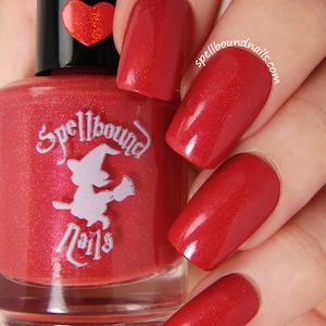 May include: A red nail polish bottle with a clear glass bottle and a black cap. The bottle has a white label with the text "Spellbound Nails" and a cartoon witch with a broom. The bottle is being held by a hand with red nail polish.