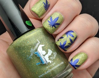 May include: A bottle of Spellbound Nails nail polish next to a hand with glittery green nail polish and blue cannabis leaf designs. The bottle is olive green with a black cap and a green heart sticker.