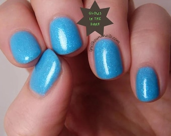 May include: Bright blue nail polish with a subtle shimmer finish applied to five fingernails. A dark star-shaped graphic is positioned between the fingers, featuring the text GLOWS IN THE DARK and the website address spellboundnails.com.
