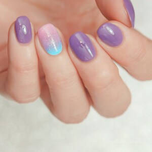 Puede incluir: Primer plano de una mano con cuatro uñas pintadas en diferentes tonos de morado y rosa. El dedo anular tiene un esmalte de uñas degradado de rosa a azul con purpurina.
