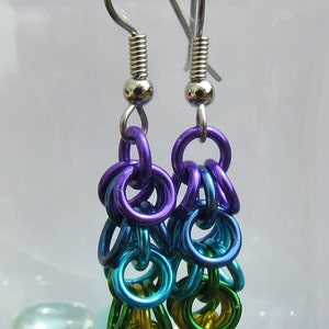 May include: A pair of chainmaille earrings. The earrings are made with a cascade of colourful metal rings in shades of purple, blue, teal, green, and yellow. The ear wires are silver-coloured.