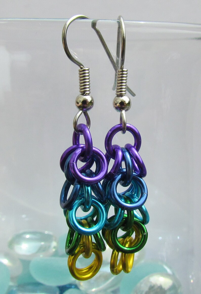 May include: A pair of chainmaille earrings. The earrings are made with a cascade of colourful metal rings in shades of purple, blue, teal, green, and yellow. The ear wires are silver-coloured.