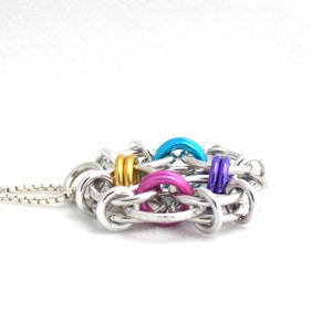 Puede incluir: Un collar de cadena de plata con un colgante colorido hecho de anillos de metal entrelazados. El colgante presenta anillos azules, rosas, morados y dorados.