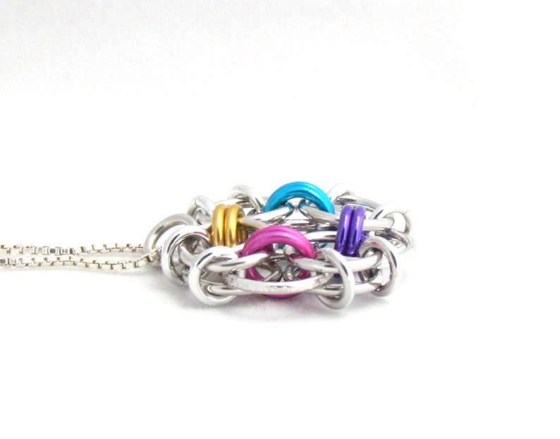 Puede incluir: Un collar de cadena de plata con un colgante colorido hecho de anillos de metal entrelazados. El colgante presenta anillos azules, rosas, morados y dorados.
