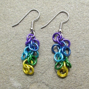 May include: A pair of handmade earrings in a rainbow colour scheme. The earrings feature a cascade of small, colourful metal rings in purple, blue, green, and gold, suspended from silver-coloured ear wires.