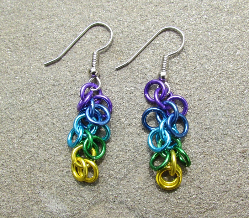 May include: A pair of handmade earrings in a rainbow colour scheme. The earrings feature a cascade of small, colourful metal rings in purple, blue, green, and gold, suspended from silver-coloured ear wires.