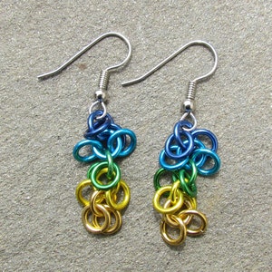 Może przedstawiać: Para kolczyków wiszących z gradientem niebieskich, zielonych i złotych metalowych kółek. Kolczyki mają srebrne haczyki.