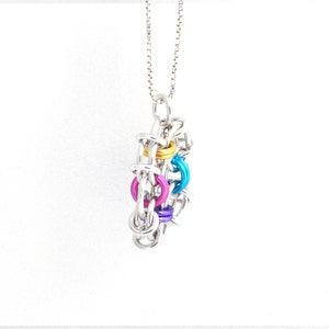 Puede incluir: Un collar de cadena de plata con un colgante colorido hecho de anillos de metal interconectados. El colgante presenta anillos en tonos de azul, rosa, morado y dorado.