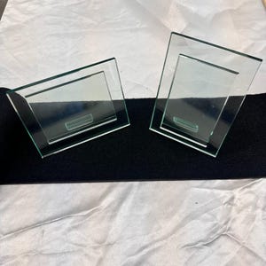 May include: Two clear glass picture frames with a simple, frameless design. One frame is positioned horizontally, while the other is oriented vertically. Both frames feature a transparent glass border and a central rectangular display area.