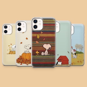 May include: Five clear smartphone cases featuring Snoopy and Woodstock in various autumn scenes. Designs include Snoopy sitting on a pumpkin, resting on a striped background, near a red doghouse, and playing in fallen leaves with trees.