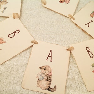 May include: A banner with the letters "B A B Y" in brown lettering on white cards. Each card features a different illustration from the Peter Rabbit series. The cards are connected with white string.