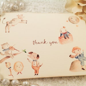 May include: A thank you card with a watercolor illustration of a cow, a cat playing a violin, a dog, a little prince, a moon, and a boy playing a horn. The card has the words "thank you" written on it.