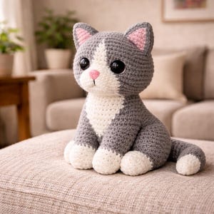May include: A handmade crochet cat doll in gray and white. The cat has large black eyes, a pink nose, and pink inner ears. The cat is sitting on a neutral-colored surface. The cat's paws and chest are white.