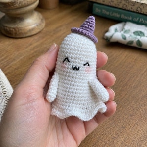 May include: A handmade white crochet ghost with a purple witch hat. The ghost has a smiling face with rosy cheeks and small arms. The ghost's body has a wavy bottom edge. A cute, whimsical Halloween decoration.