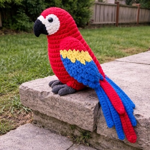 May include: A handmade crochet parrot in vibrant red, yellow, and blue. The bird has a black beak, gray feet, and a white eye. The parrot is sitting on a stone surface, showcasing its detailed craftsmanship.