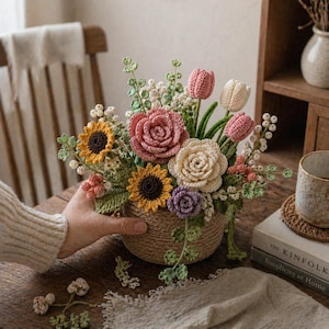 Può includere: Una composizione floreale all'uncinetto fatta a mano in un cestino intrecciato. La composizione presenta girasoli, rose, tulipani e altri fiori nei toni del rosa, giallo e crema. Il cestino è tenuto dalla mano di una persona e la composizione è su un tavolo di legno.