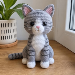 May include: A crocheted cat toy in shades of grey and white, featuring black eyes and a pink nose. The cat has grey stripes and pink inner ears. It is sitting on a wooden surface. The cat's tail is curled to the side.