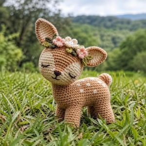 Könnte beinhalten: Gehäkelte Hirschfigur, braun mit weißen Punkten, mit einem Blumenschmuck aus rosa und weißen Blumen. Der Hirsch hat große Ohren und geschlossene Augen. Er steht im grünen Gras.