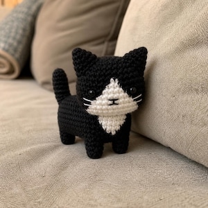 Könnte beinhalten: Eine handgefertigte Häkelkatzenfigur in Schwarz und Weiß. Die Katze hat ein weißes Gesicht und eine weiße Brust, mit schwarzen Ohren und Körper. Sie hat kleine, schwarze Augen und weiße Schnurrhaare. Die Katze steht auf einer neutralfarbenen Oberfläche.
